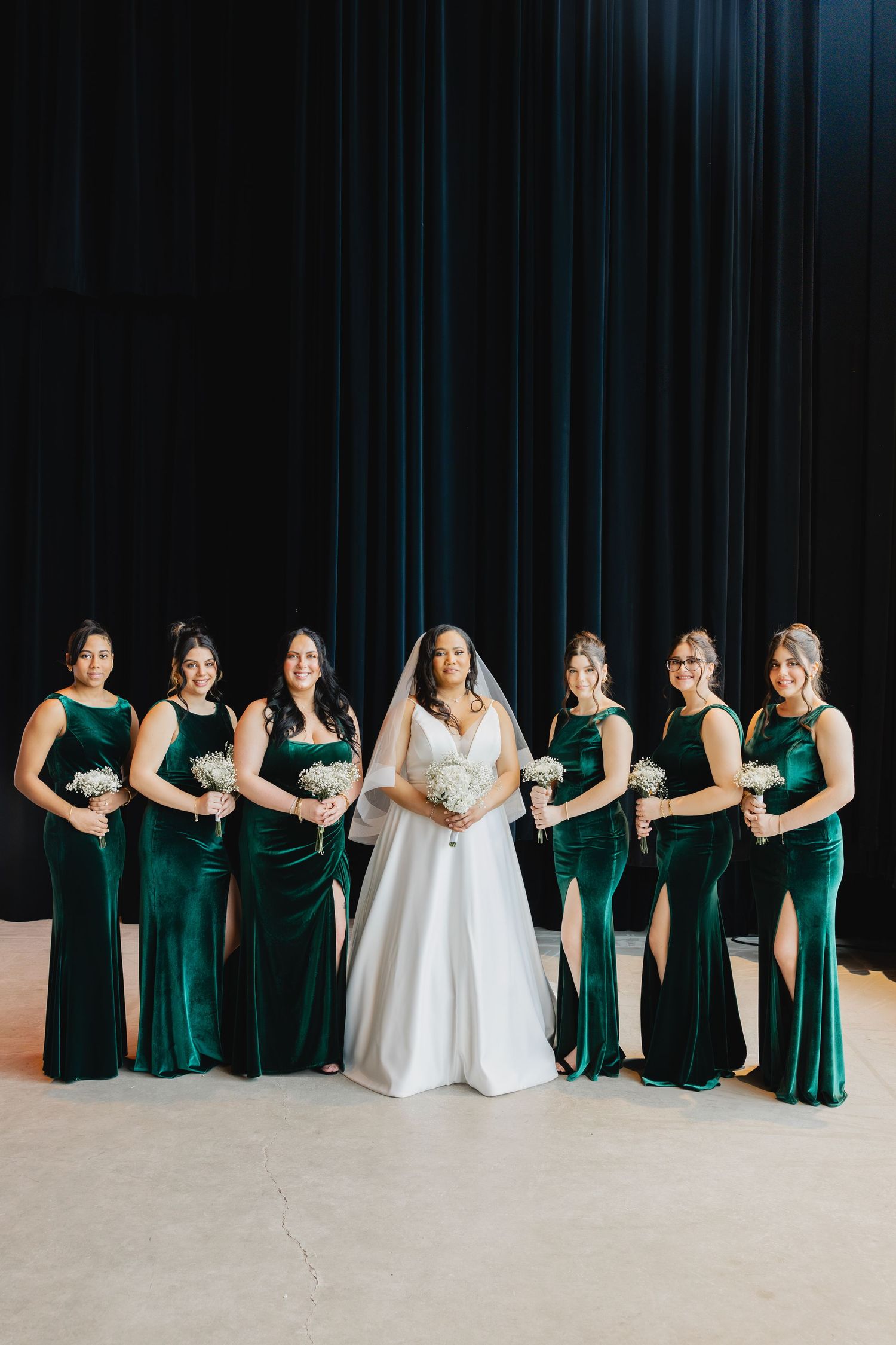 Bride in white gown poses with six bridesmaids in emerald green velvet dresses holding white bouquets.