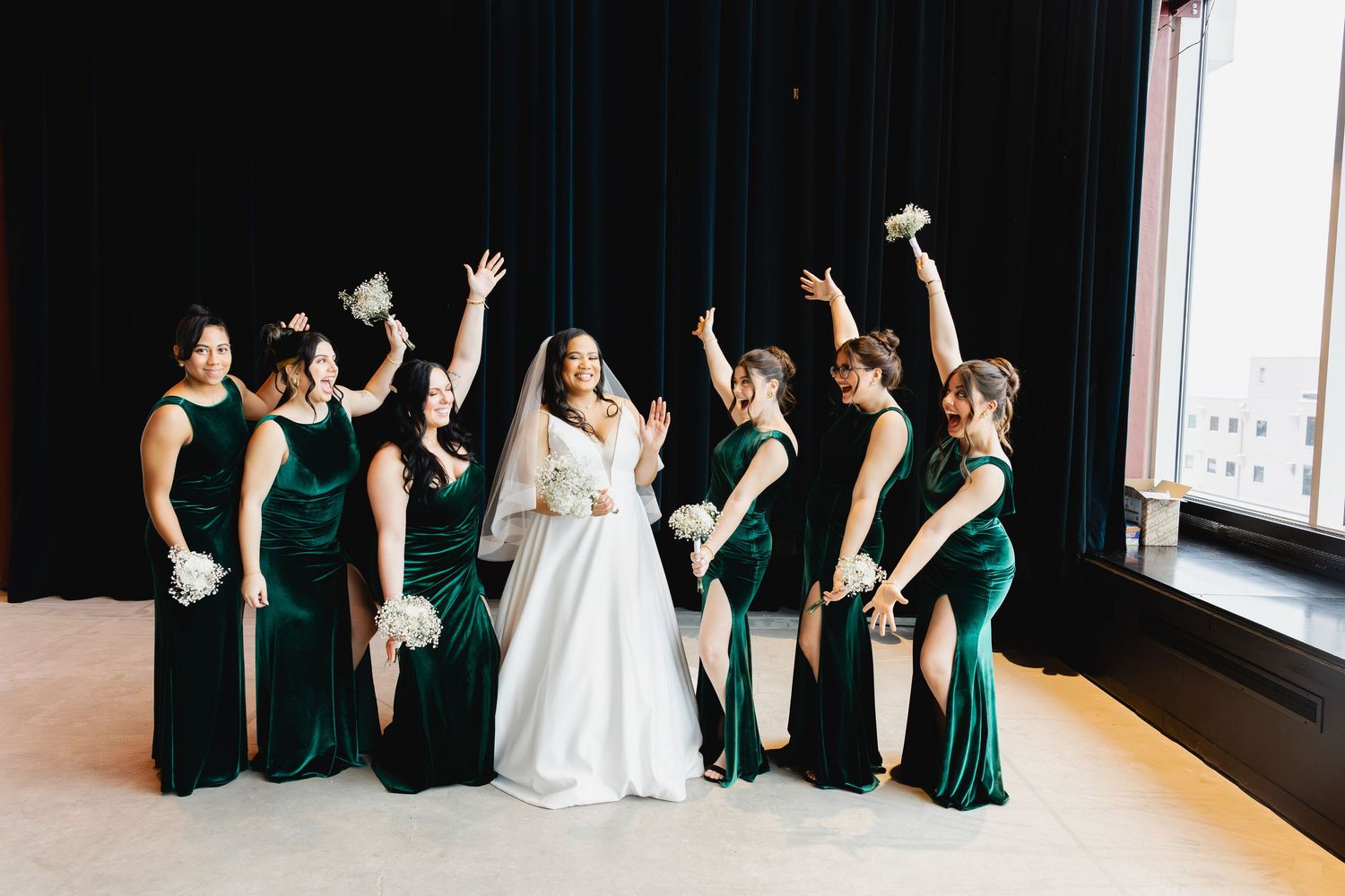Bride in white gown poses with bridesmaids in emerald green velvet dresses, all celebrating with raised arms.