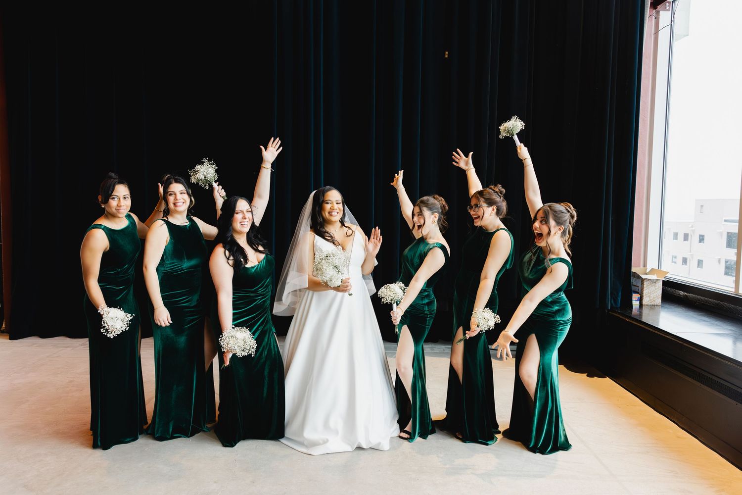 Bride in white gown poses with bridesmaids in emerald green velvet dresses, holding bouquets and cheering joyfully.