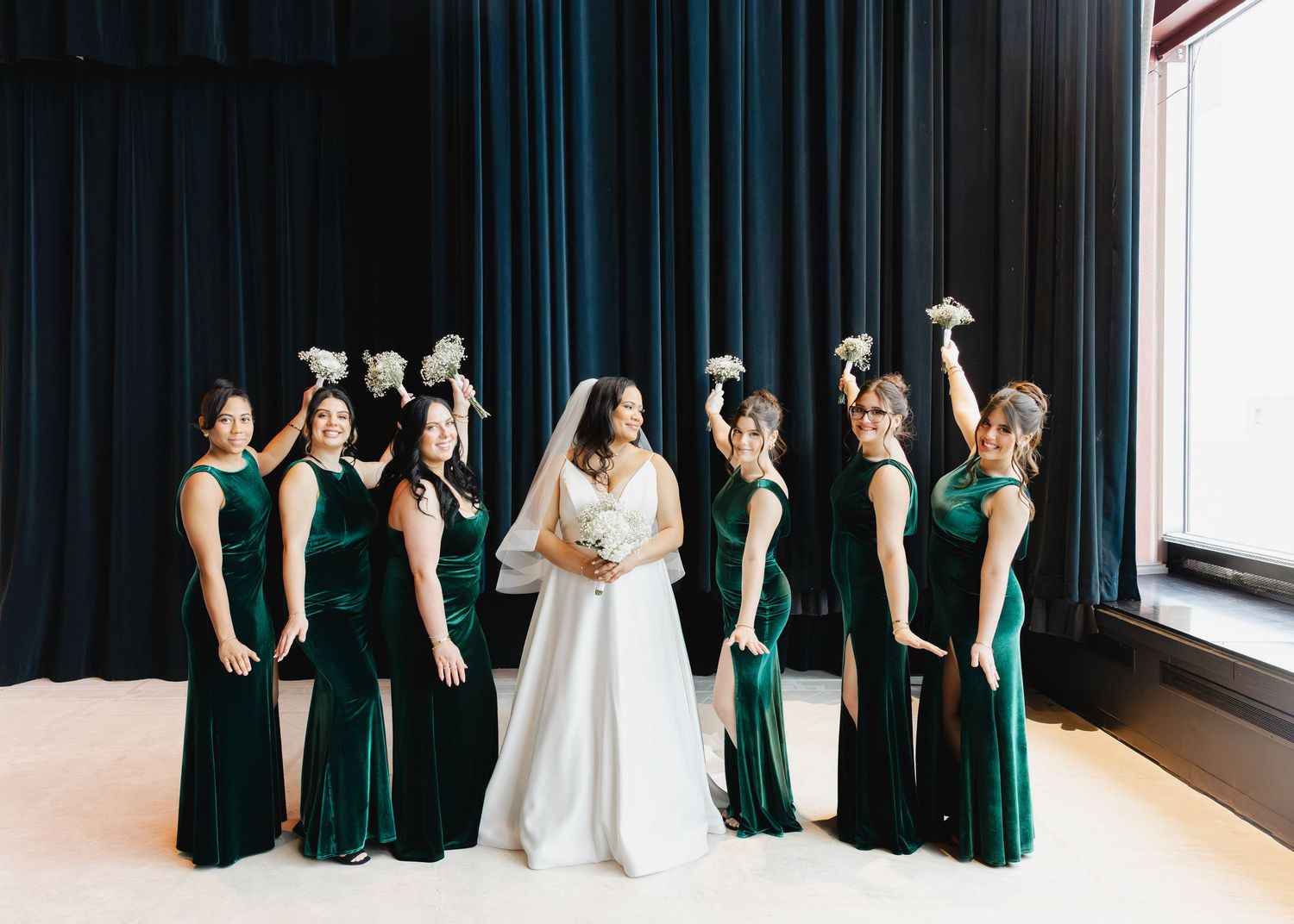 Bride in white gown poses with six bridesmaids in emerald green velvet dresses holding white bouquets against dark curtains.
