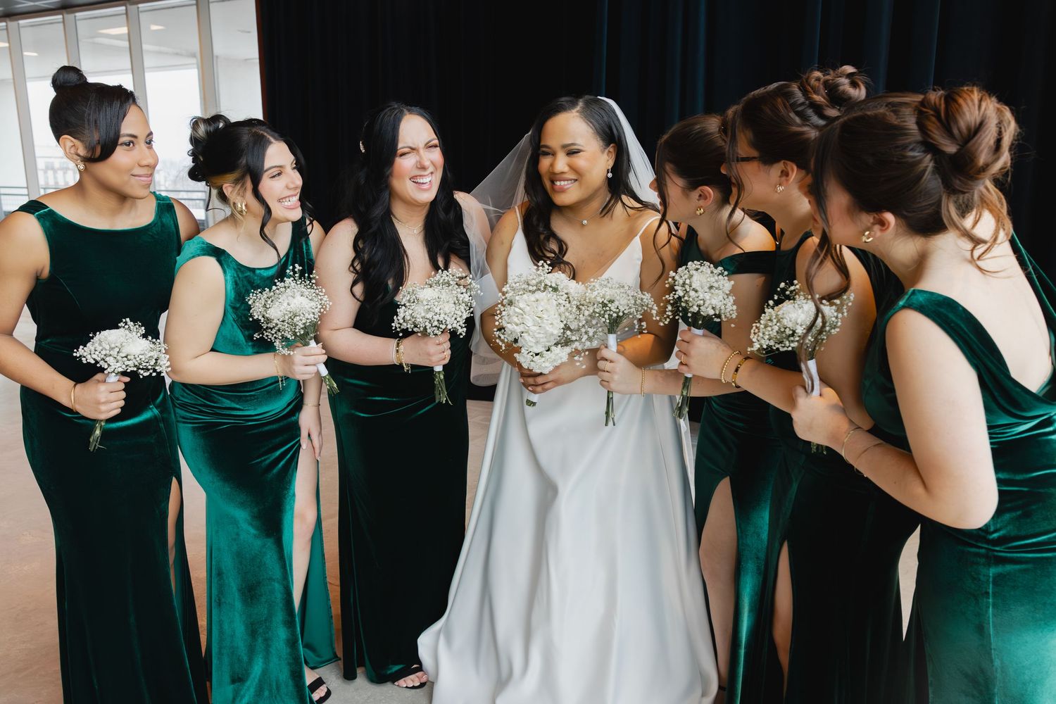 Bride in white gown with bridesmaids in emerald green velvet dresses holding white bouquets indoors.