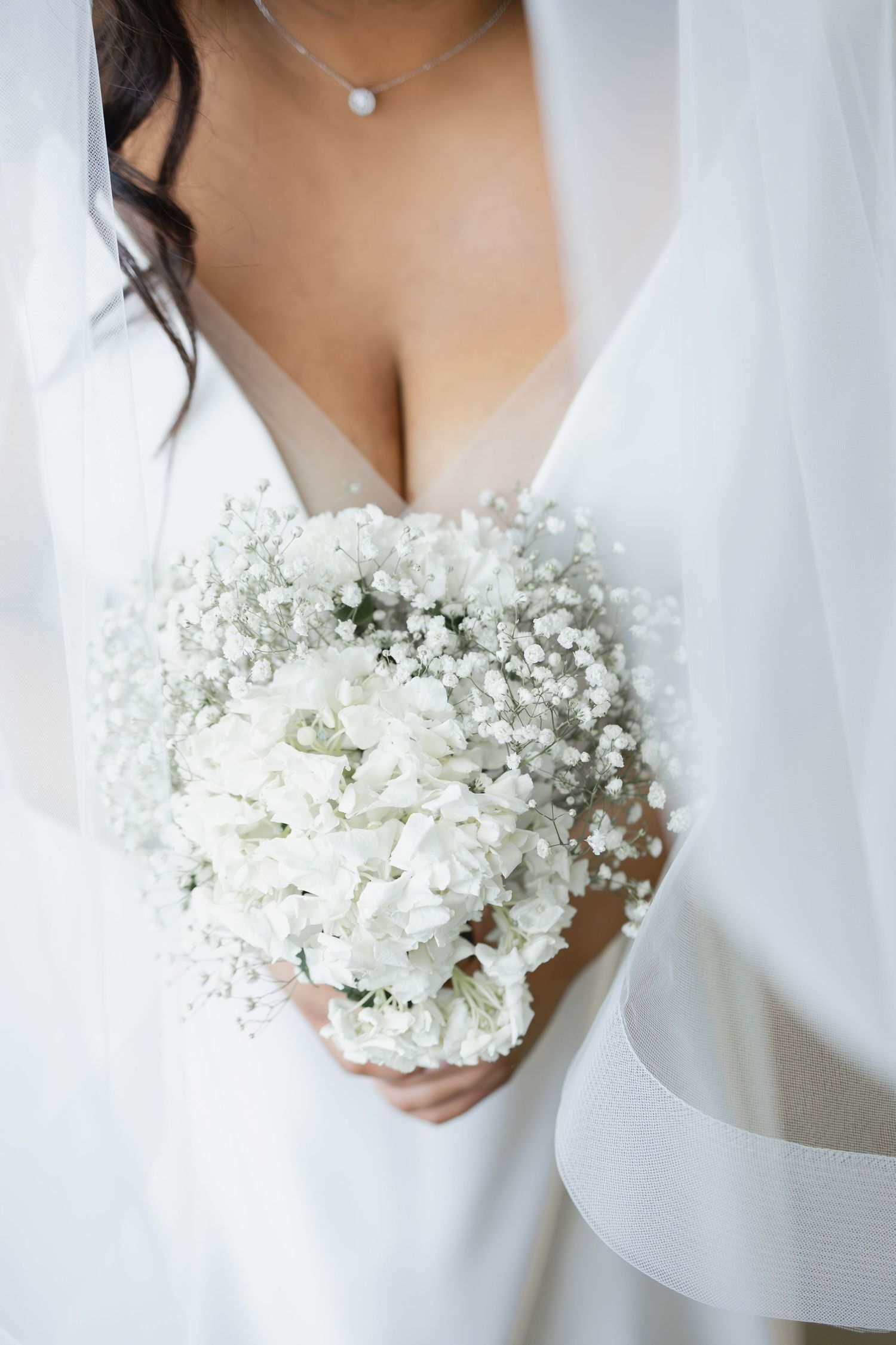 Bride in white gown holding elegant all-white bouquet of baby's breath and roses, wearing delicate pearl necklace.