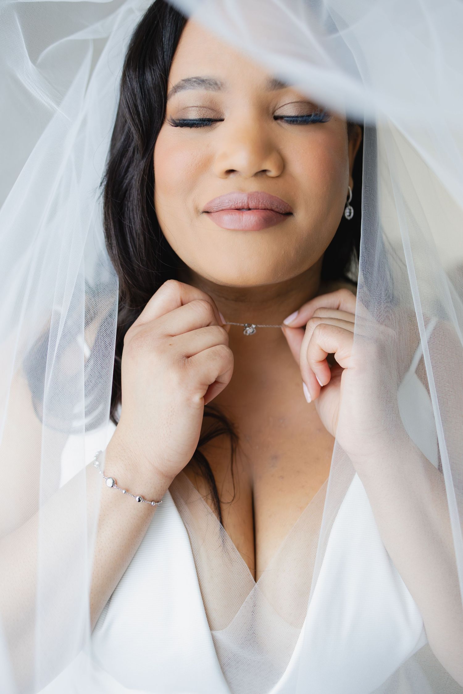 Beautiful bride with closed eyes adjusts her delicate necklace beneath her wedding veil.