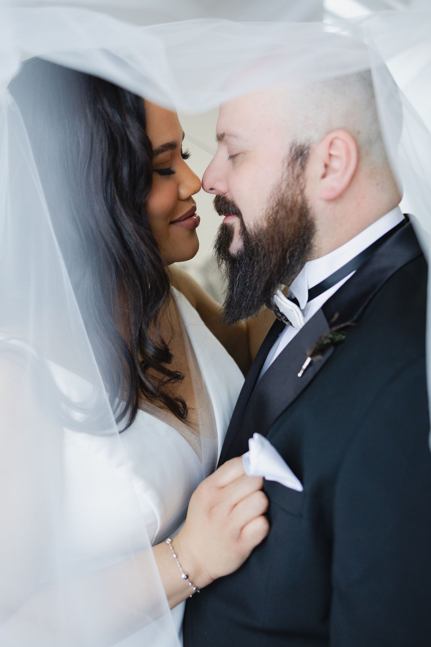 Bride and groom share an intimate moment under a flowing white veil, nearly kissing in wedding attire.