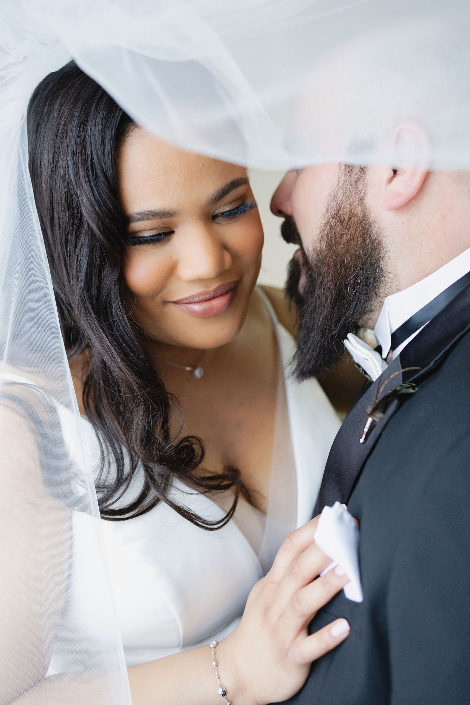 Bride and groom share an intimate moment under a wedding veil, bride smiling softly in white dress.