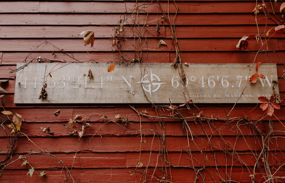 Weathered wooden sign with GPS coordinates and compass rose mounted on red barn wall with autumn vines.