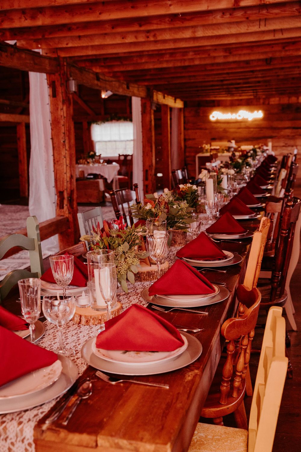 Rustic barn wedding reception with long wooden table set with red napkins, crystal glasses, and floral centerpieces under warm lighting.