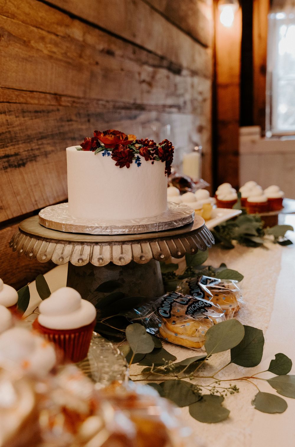 White single-tier wedding cake with red florals on a wooden cake stand surrounded by cupcakes and greenery on a dessert table.