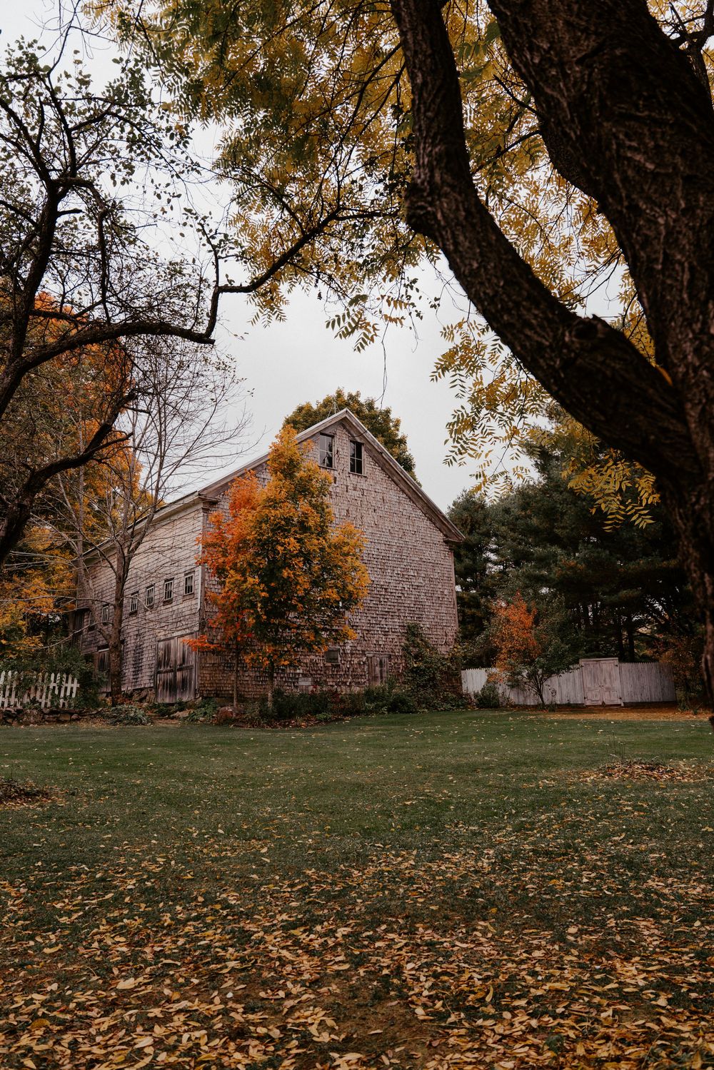 Old stone barn surrounded by autumn foliage with fallen leaves on green grass under overcast skies.