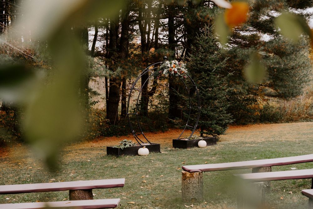 Outdoor wedding ceremony seating with wooden benches on grass, fire pit, hay bales, and tall evergreen trees in autumn.