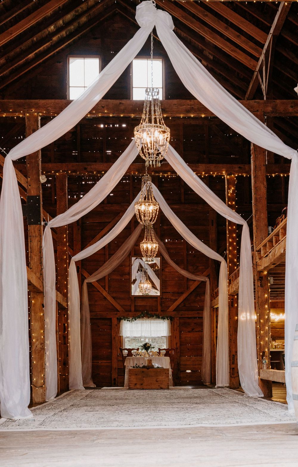 Rustic barn wedding venue with white draping, crystal chandeliers, string lights, and elegant altar setup.