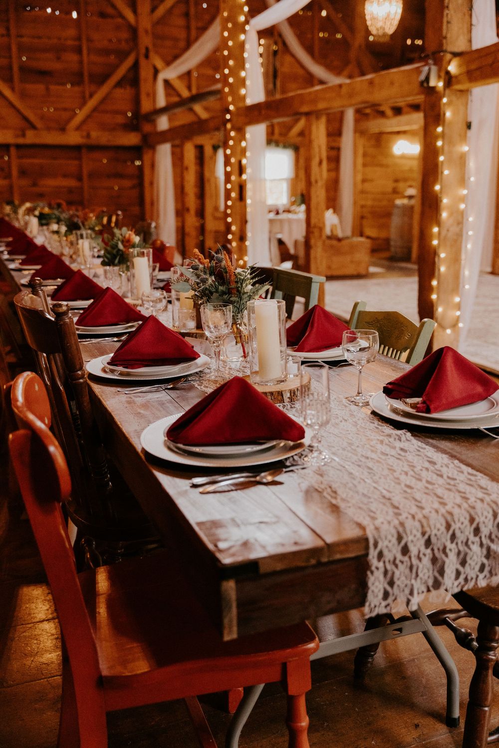 Rustic barn wedding reception with long wooden tables, red napkins, lace runners, candles, and string lights.