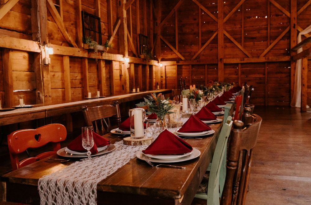 Rustic barn wedding reception with long wooden tables, red napkins, lace runner, candles, and string lights in warm wood interior.