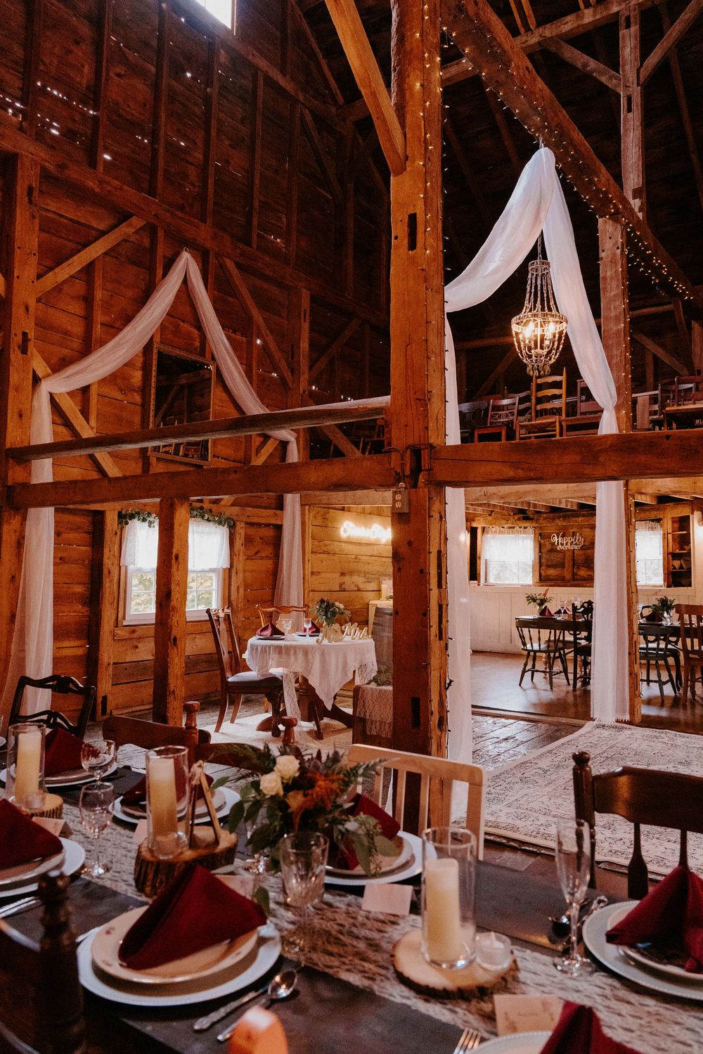 Rustic barn wedding venue with elegant table settings, burgundy napkins, floral centerpieces, string lights, and draped white fabric decor.