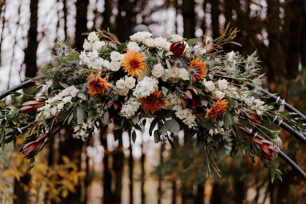 Rustic wedding arch adorned with white, orange dahlias, greenery, and protea flowers in a forest setting.