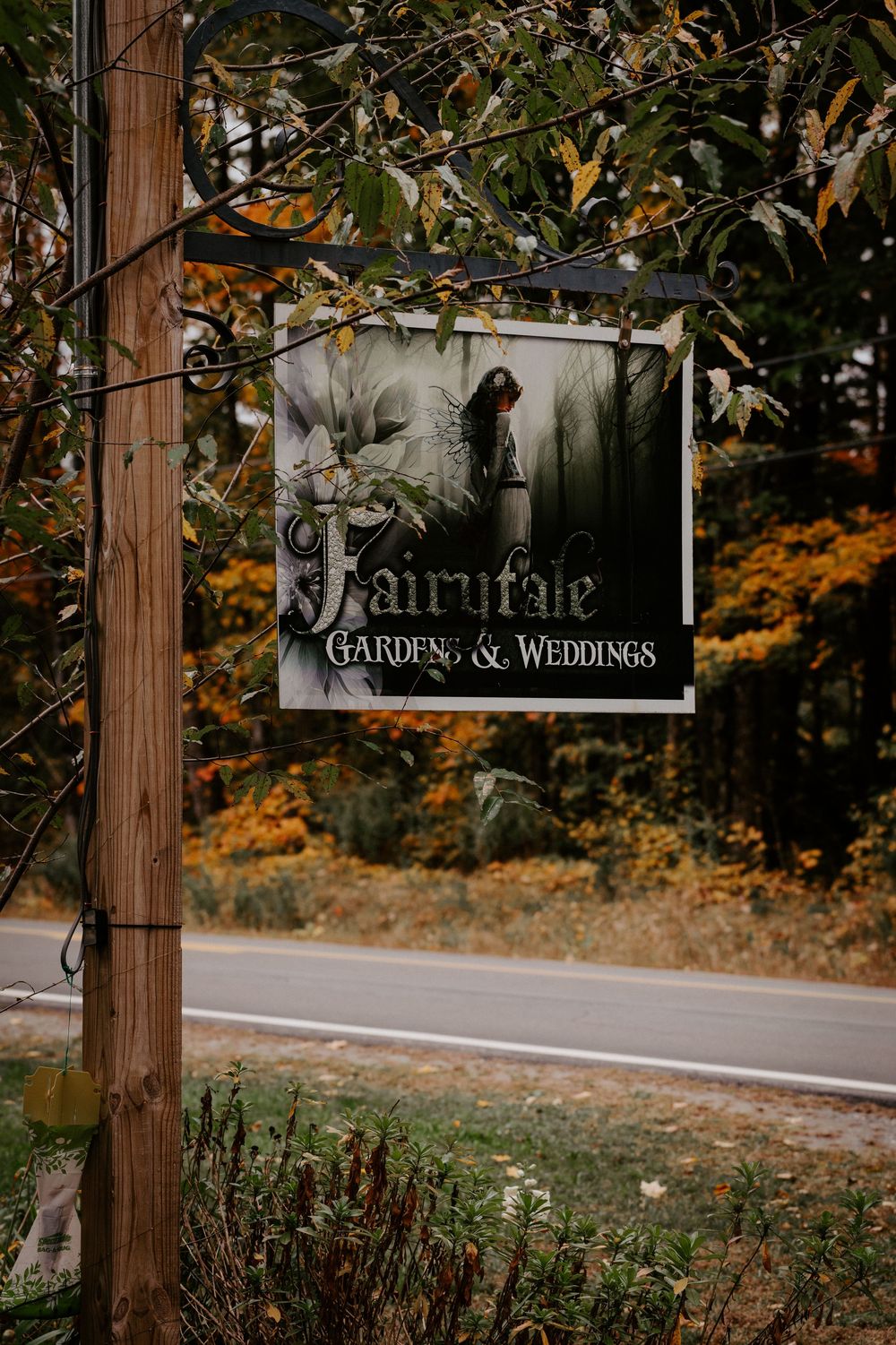 Fairytale Gardens & Weddings sign on a wooden pole surrounded by autumn foliage along a scenic road.