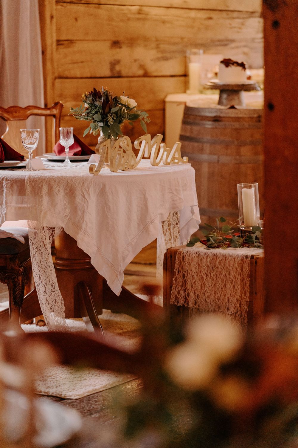Rustic barn wedding reception with lace-draped Mr. & Mrs. sweetheart table, floral centerpiece, wine glasses, and wooden barrel backdrop.
