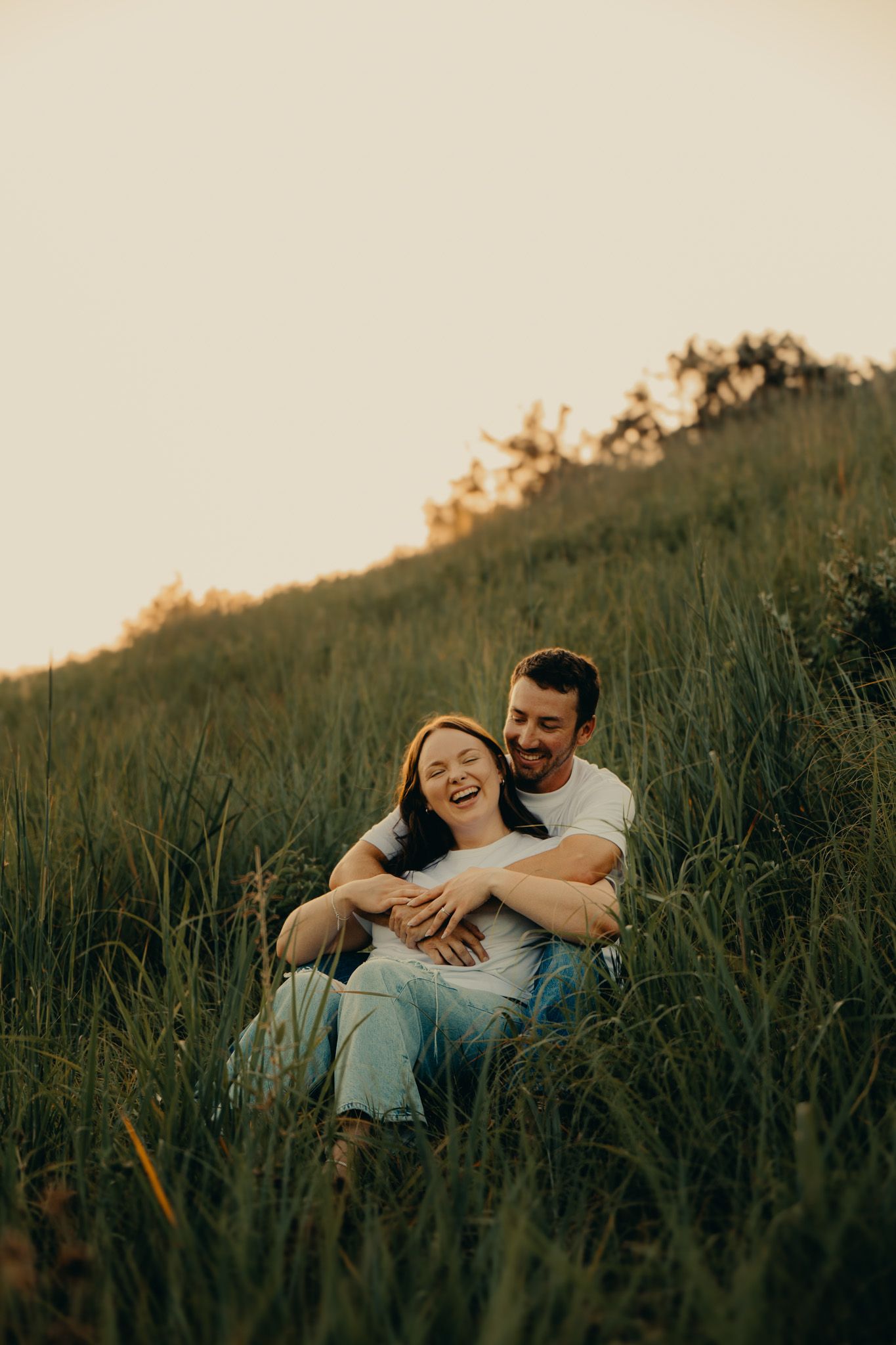 A couple laughs and embraces while sitting in tall green grass on a hillside during golden hour.