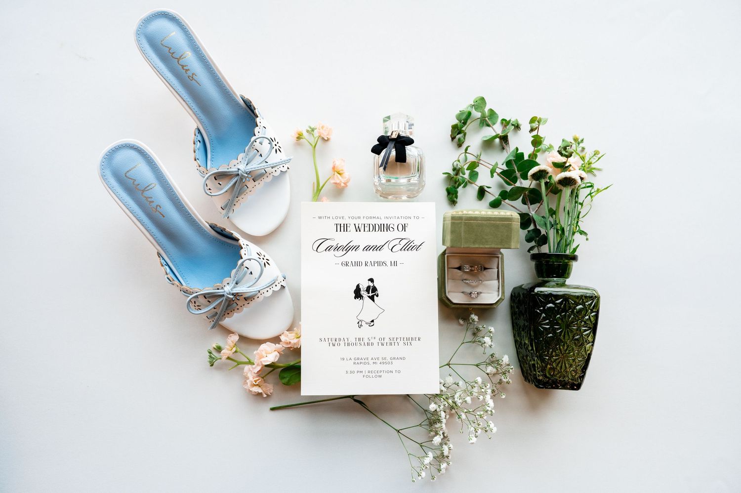 Elegant wedding flat lay featuring blue heels, floral invitation suite, delicate flowers, and a ring box on white background.