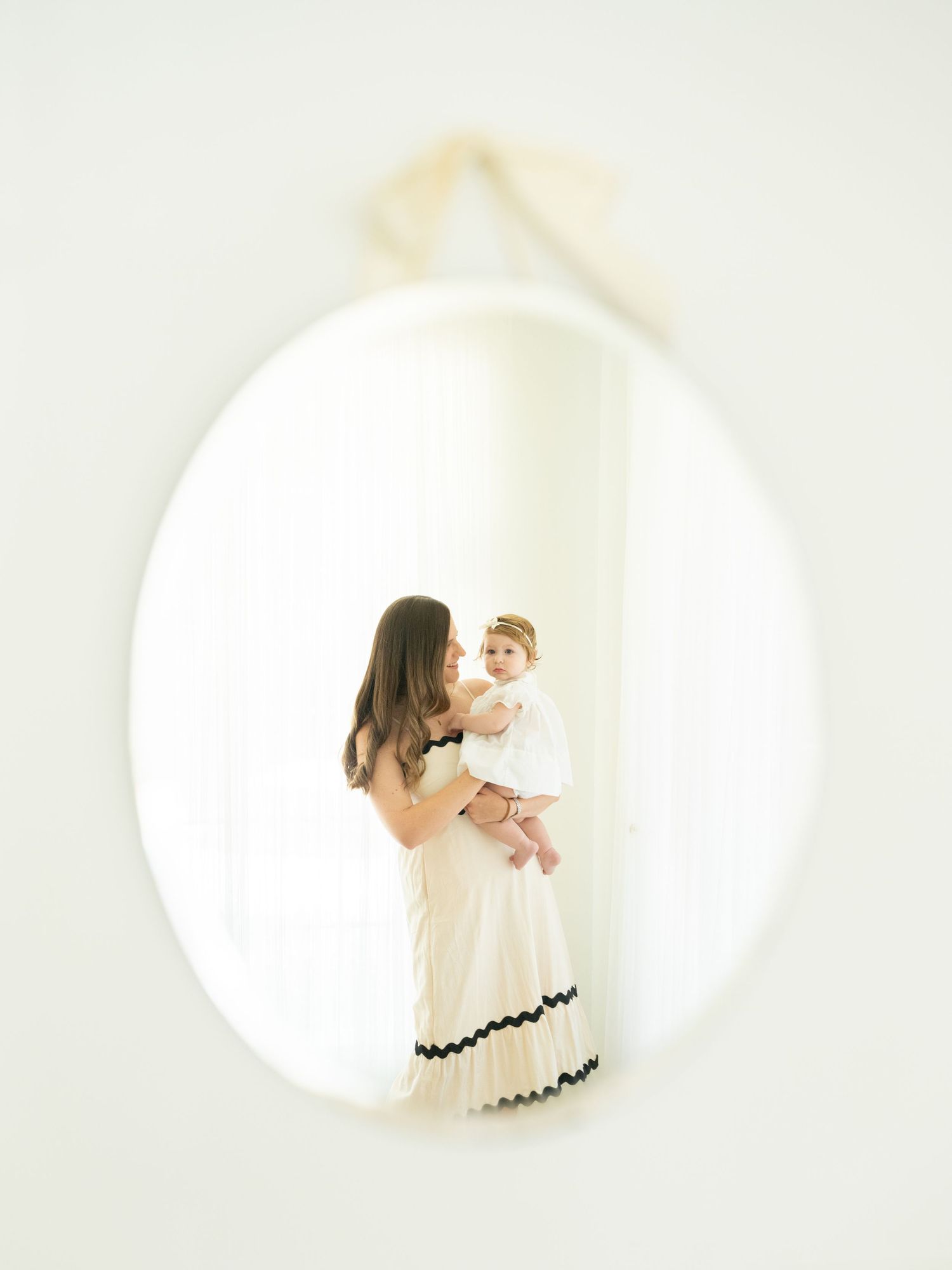 A mother holds her baby reflected in an oval mirror, both dressed in white, in a bright minimalist room.