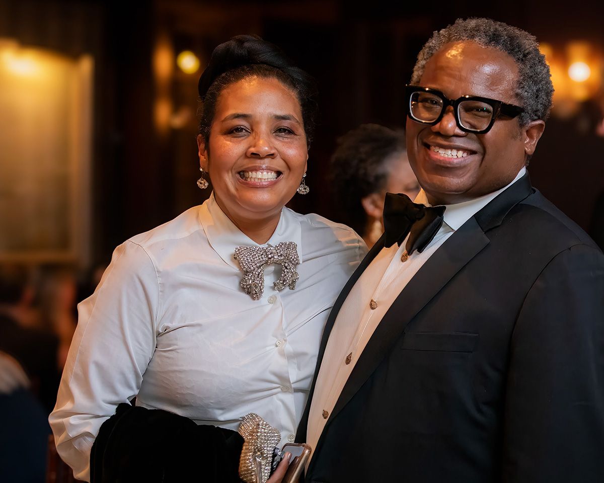 Two elegantly dressed people smiling at a formal event, wearing black and white attire with decorative accessories.
