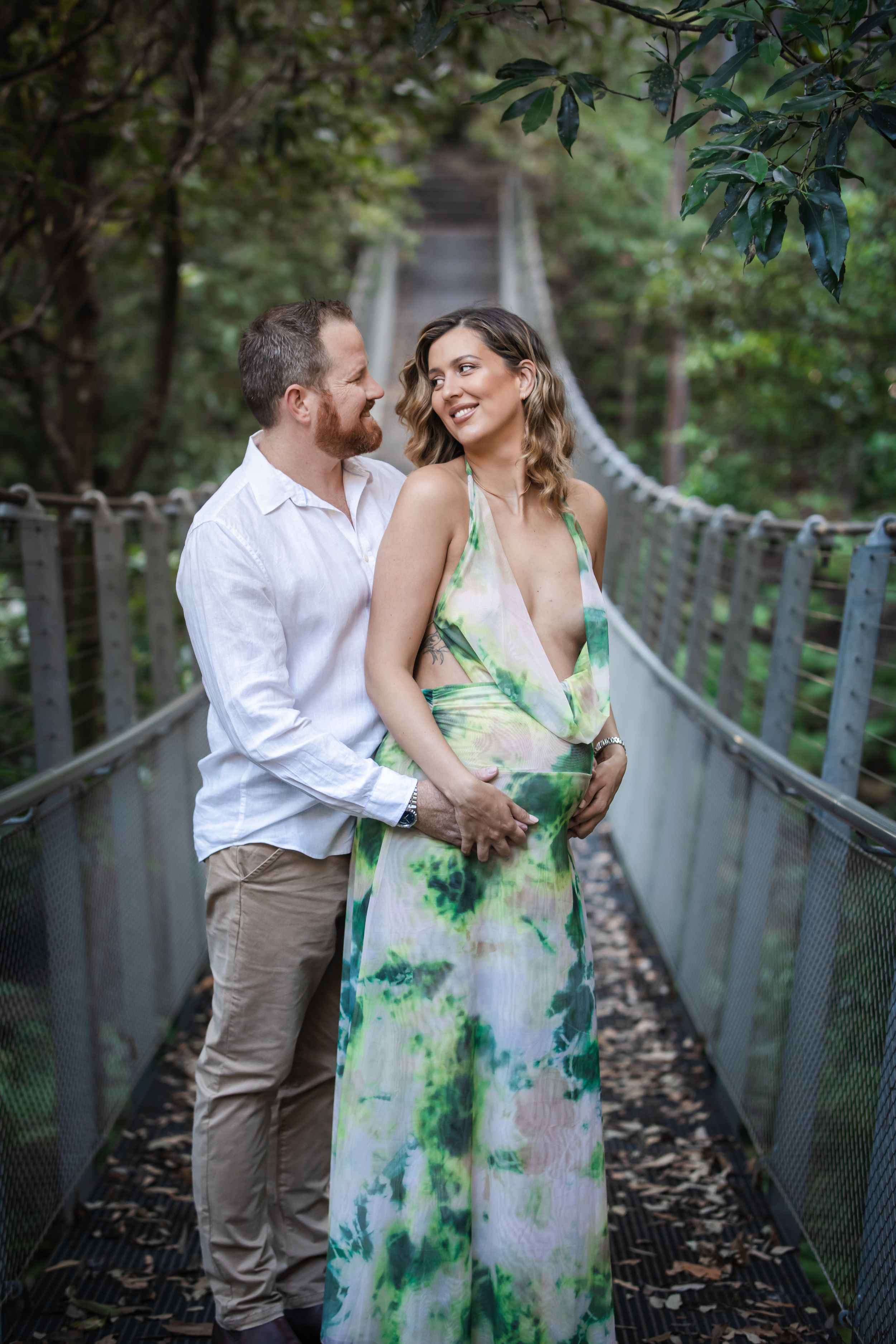 Pregnant couple embracing on a The Swing bridge in Cherrybrook in lush greenery, woman in floral dress, man in white shirt.