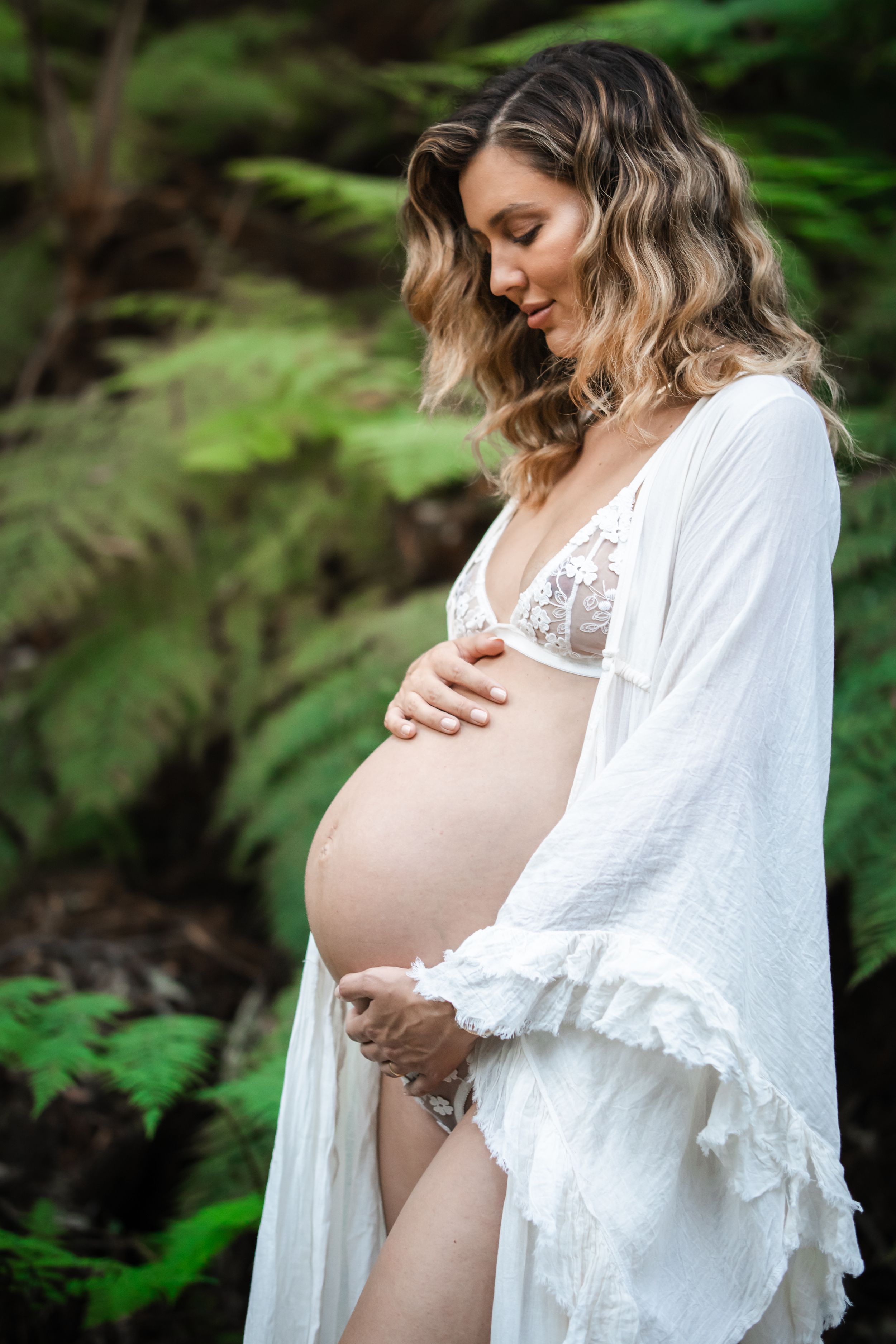 Pregnant woman in white lace lingerie and robe cradling her baby bump outdoors in Cherrybrook among lush green ferns.