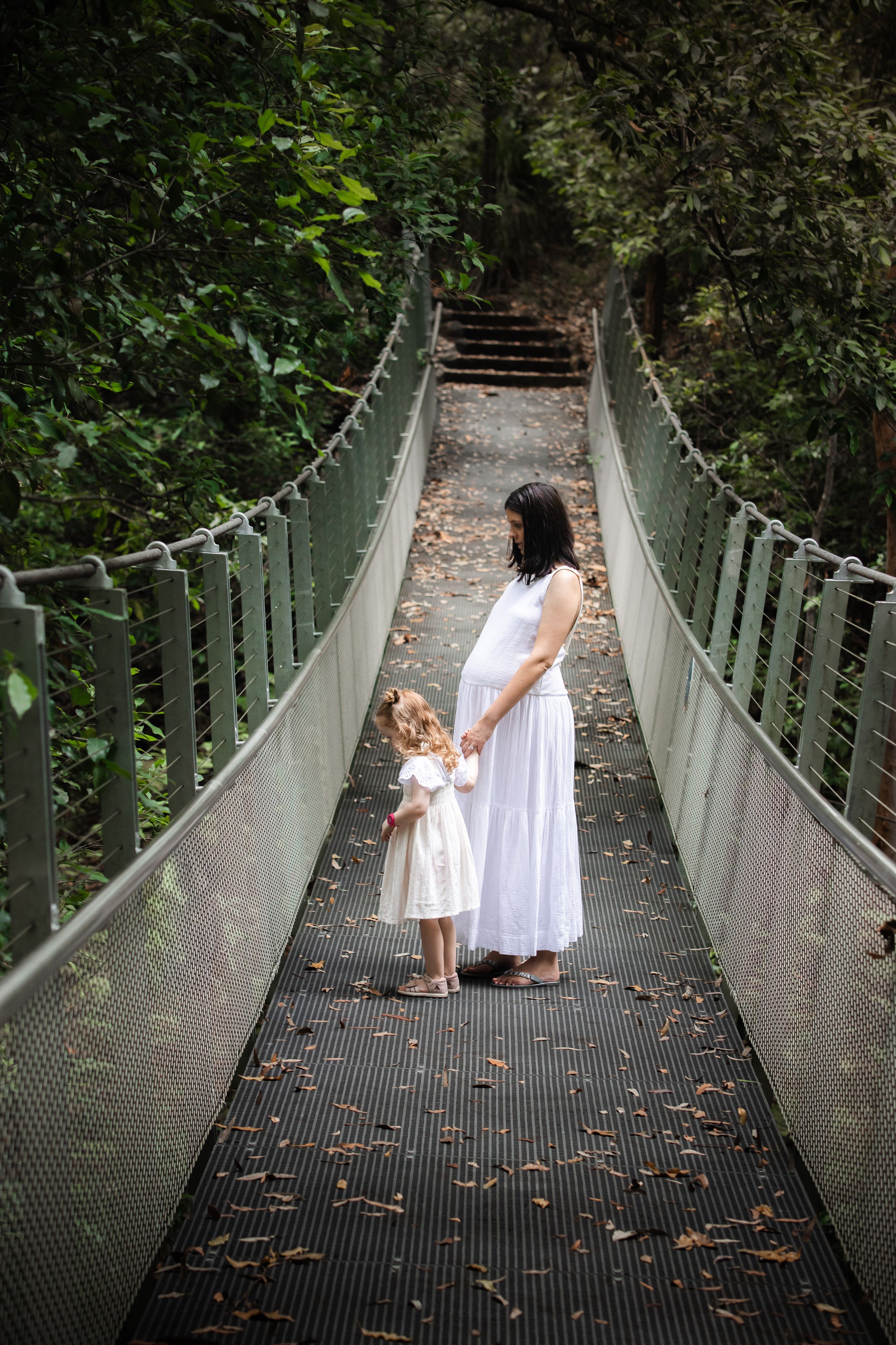 Pregnant woman in white dress with young daughter on The Swing bridge in Cherrybrook surrounded by lush green forest.
