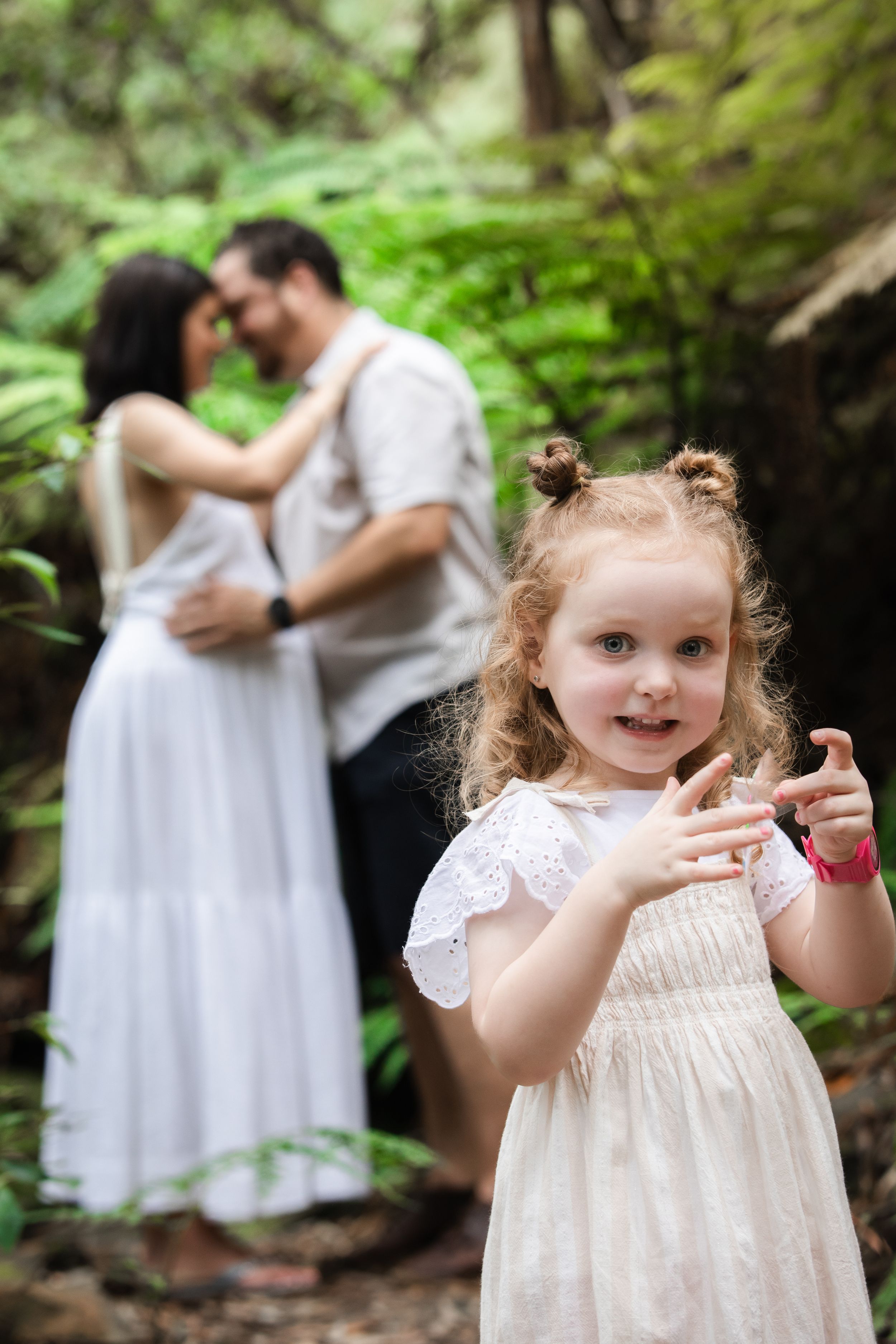 A young girl in a white dress smiles in a lush forest in Cherrybrook while her parents embrace romantically in the background.