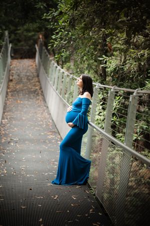 Pregnant woman in teal off-shoulder gown poses on The Swing bridge in Cherrybrook surrounded by lush greenery in a maternity photo.