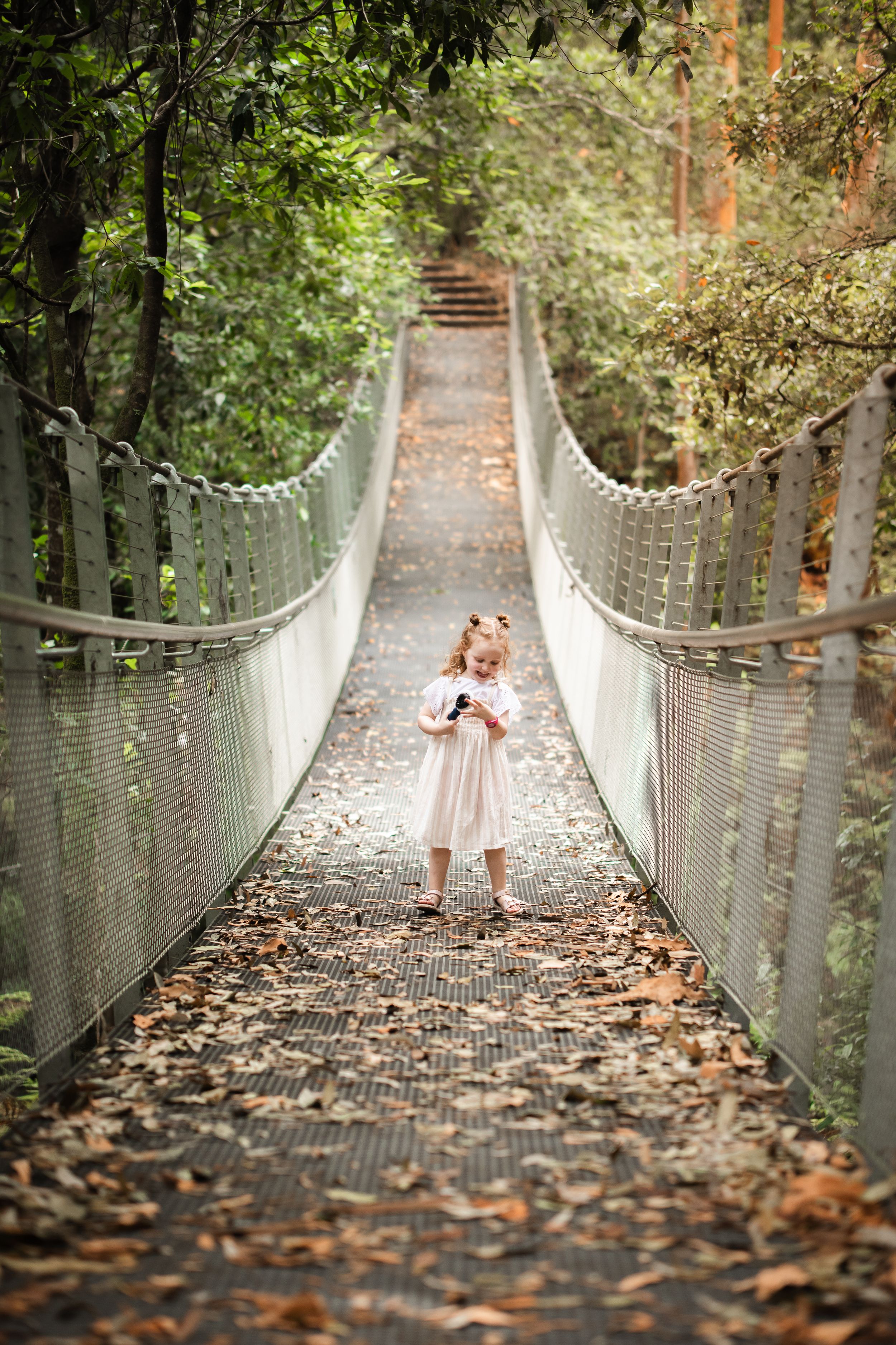 Young girl in white dress standing on The Swing bridge in Cherrybrook surrounded by lush green trees and fallen leaves.