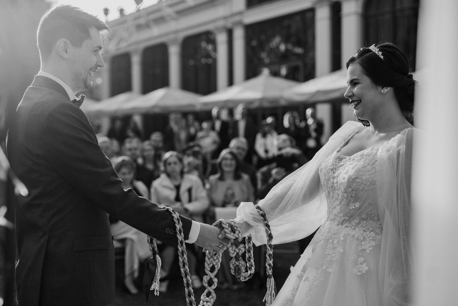 Black and white wedding photo of bride and groom exchanging vows with decorative rope during outdoor ceremony.