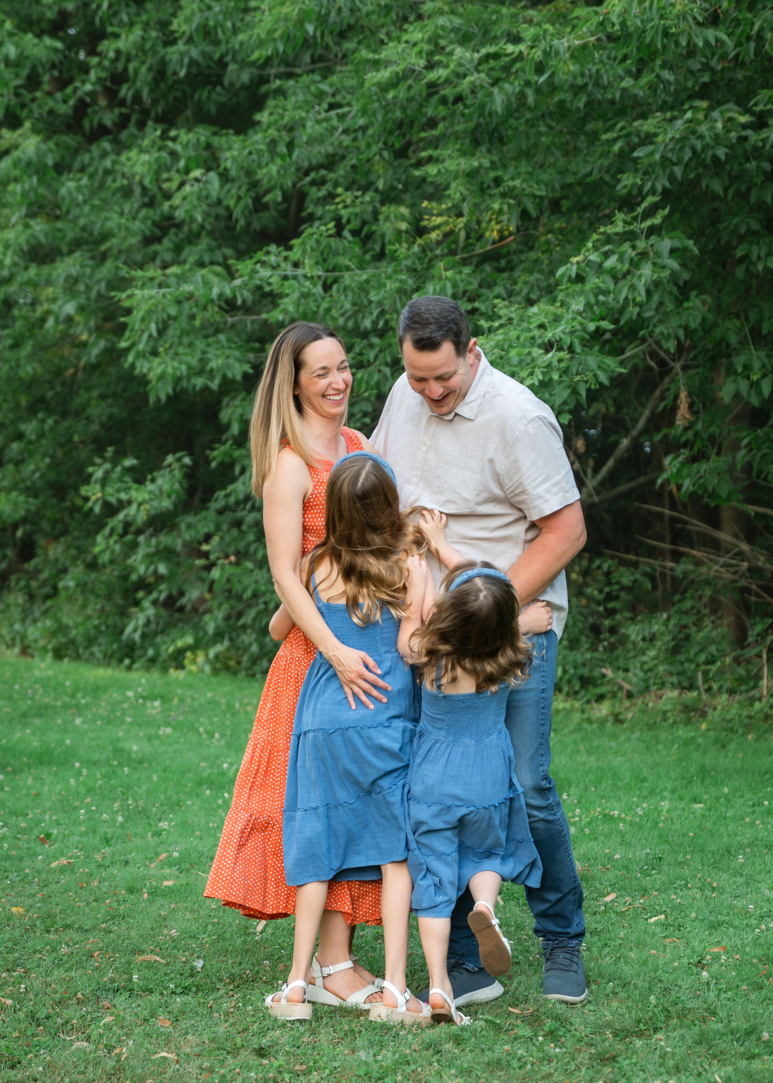 Family of four laughing together on green grass with lush trees, two girls in blue dresses hugging parents outdoors.
