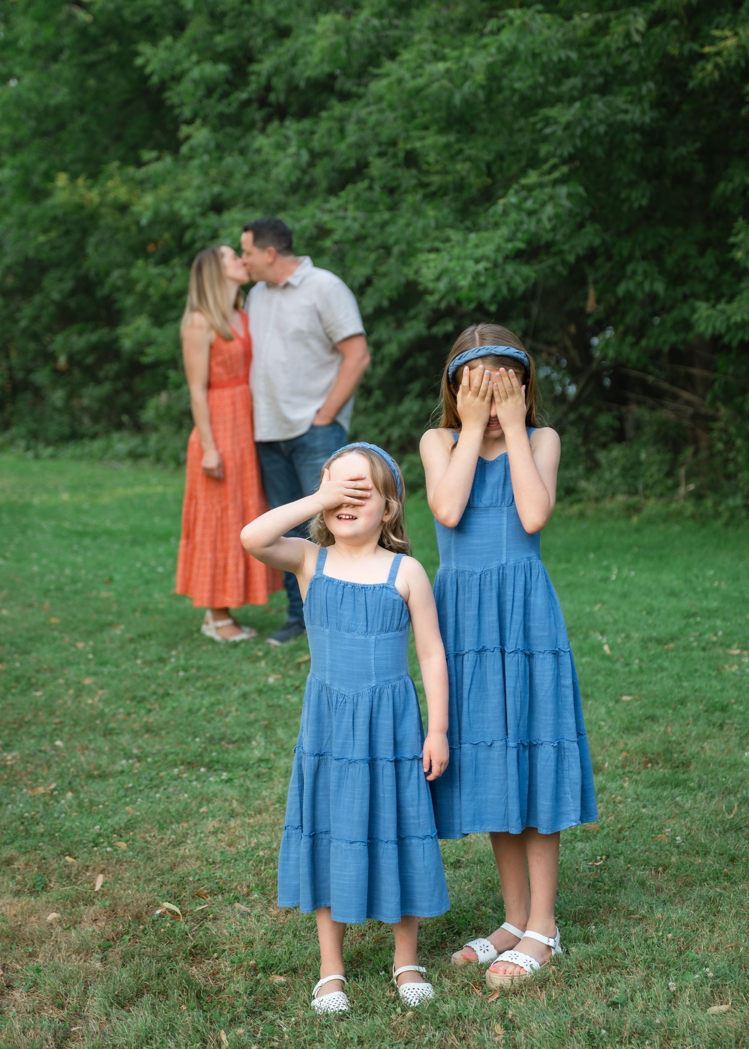 Two girls in blue dresses cover their eyes while their parents share a kiss in a green outdoor setting.
