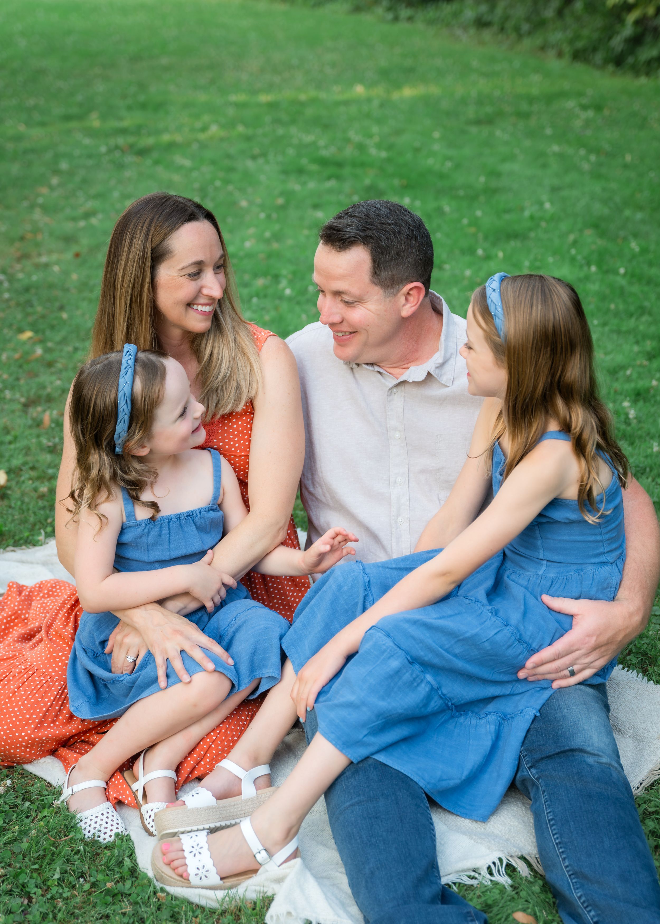 A happy family of four sits on a blanket in a park, with two girls in denim dresses and parents in red and white.