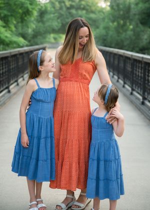 Pregnant woman in orange dress with two girls in blue dresses on a bridge.