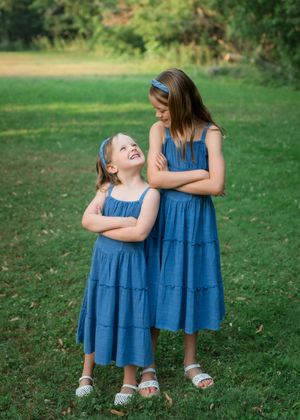 Two girls in matching blue tiered dresses smiling at each other on a green lawn.