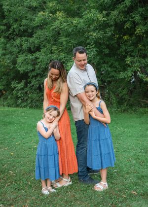Family of four posing outdoors on green grass, two girls in blue dresses with parents in orange and gray outfits.