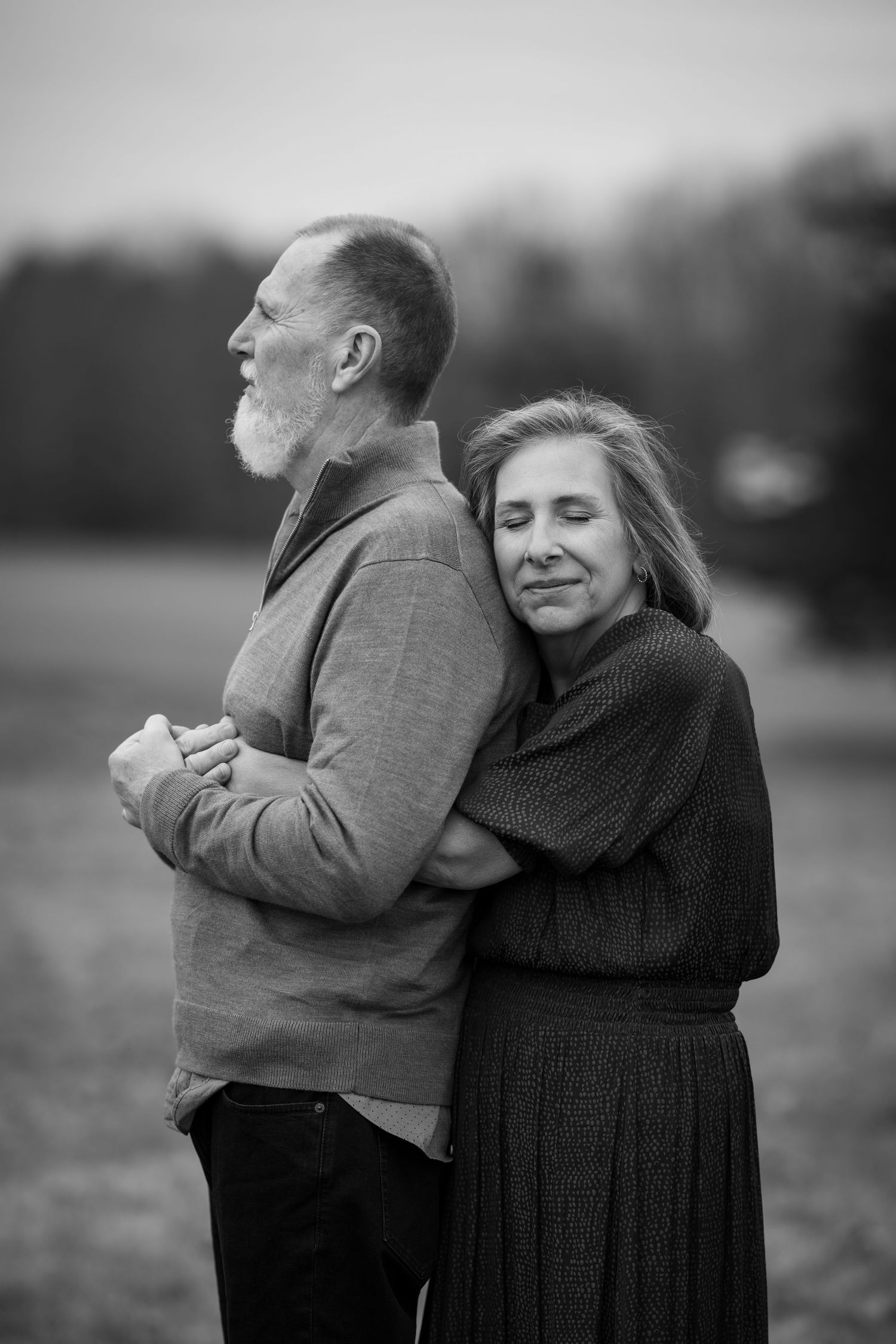 A loving black and white portrait of an older couple embracing outdoors, with the woman hugging the man from behind.