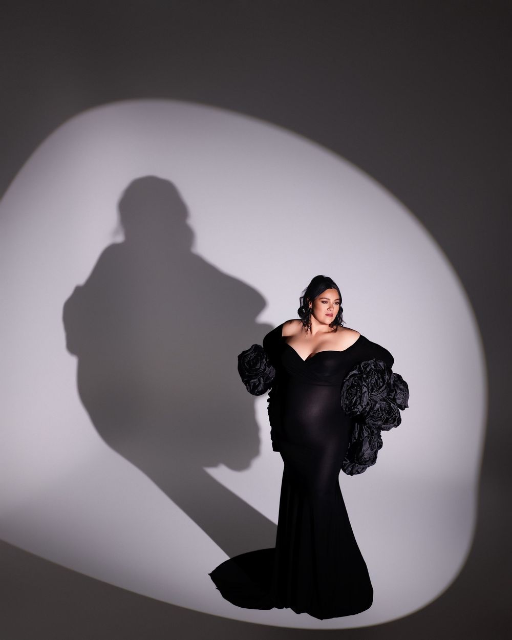 Woman in elegant black off-shoulder gown with ruffled sleeves poses dramatically against a white spotlight backdrop.