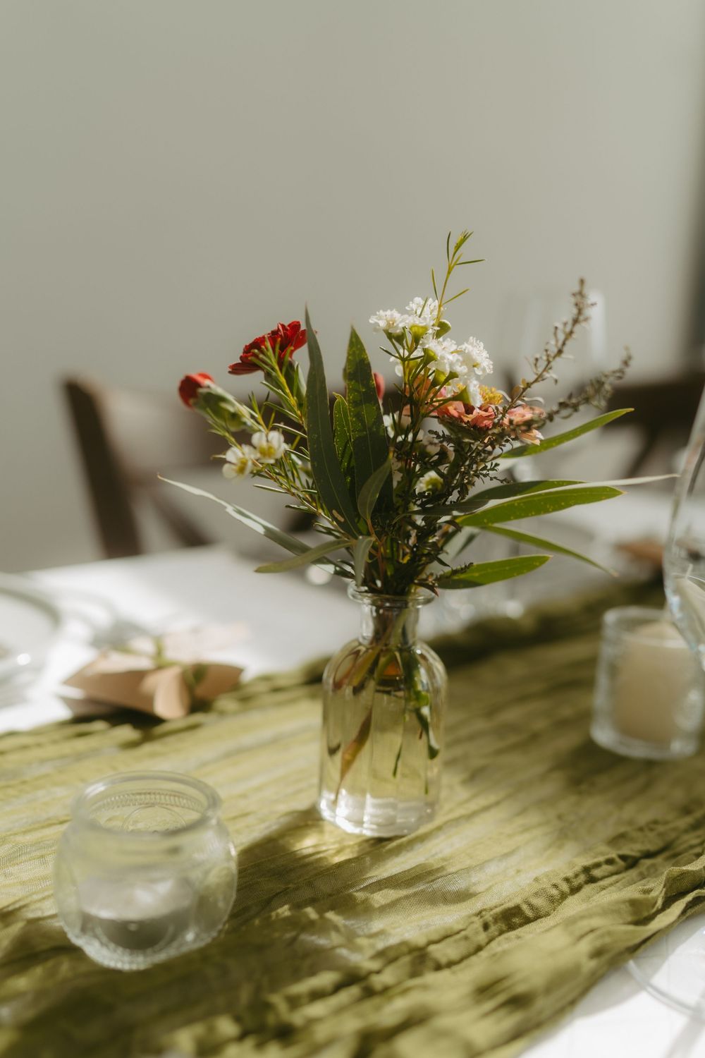 Small glass vase with wildflowers and greenery on a green table runner with candles in natural light.