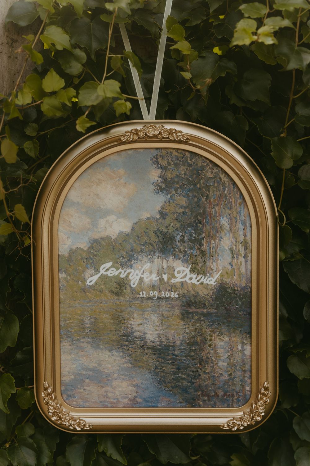 Elegant gold arched frame with Monet painting displaying Jennifer and David wedding date signage amid green ivy.