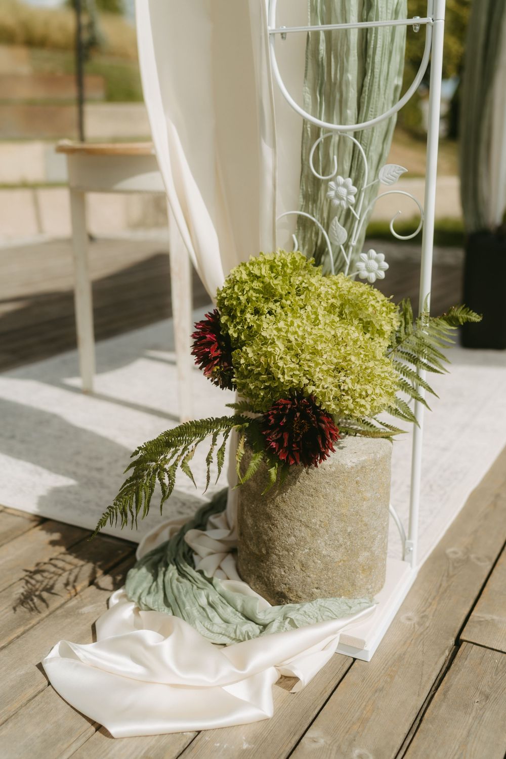 Elegant wedding ceremony decor with green hydrangeas and burgundy flowers in a stone vase beside a white ornate arch.