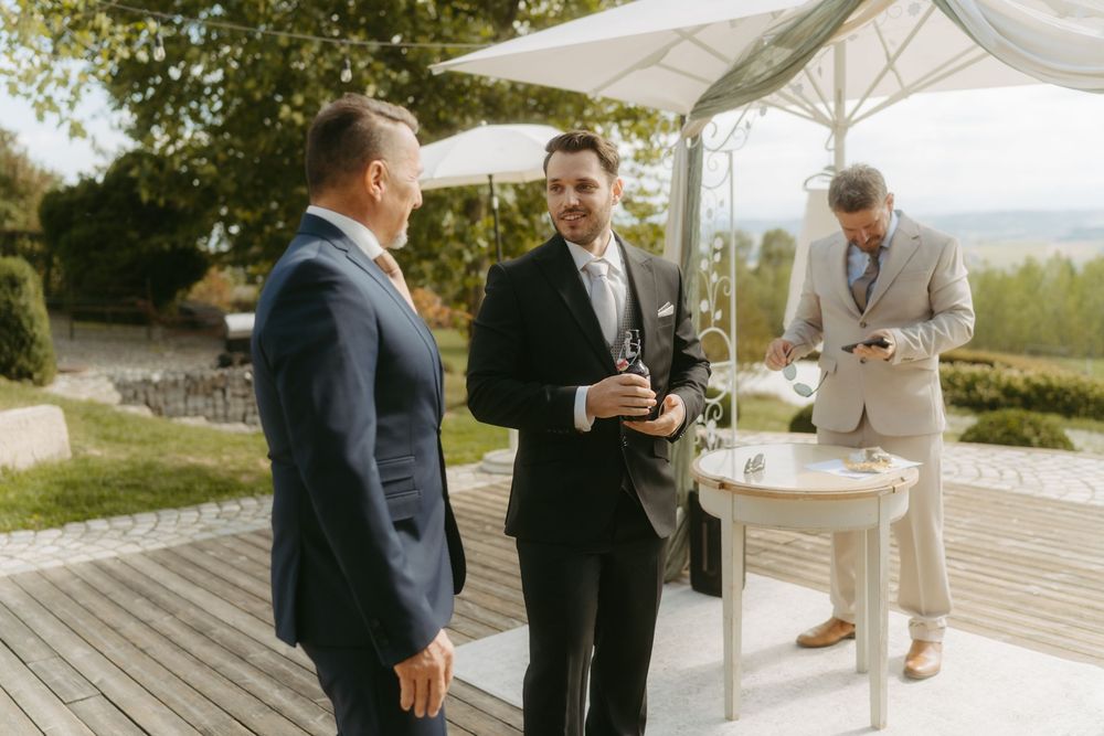 Three men in suits socializing outdoors at a wedding reception on a wooden deck with white umbrellas.