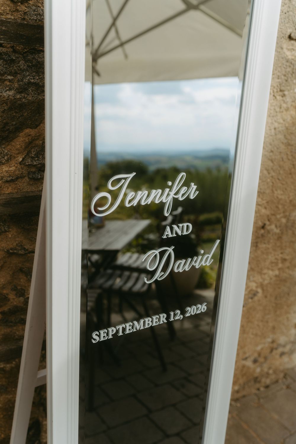 Elegant wedding mirror sign with white script reading Jennifer and David, September 12, 2026, outdoors.