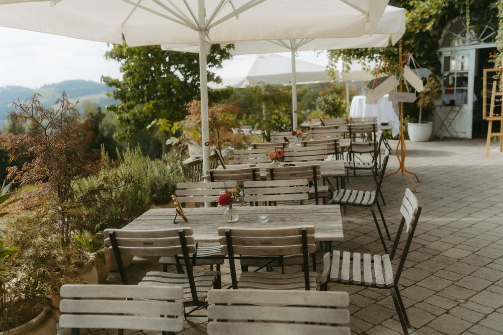 Rustic outdoor wedding reception terrace with wooden tables, chairs, and white umbrellas overlooking lush greenery.