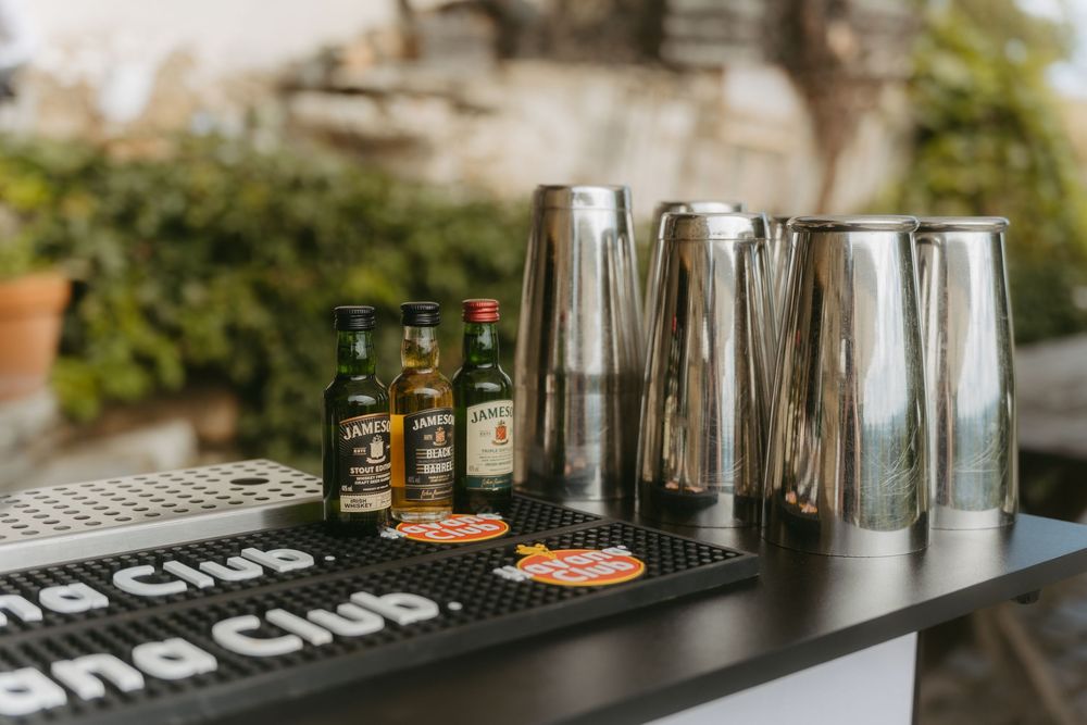 Bar setup with Havana Club mini bottles and silver cocktail shakers on a black mat at outdoor wedding.