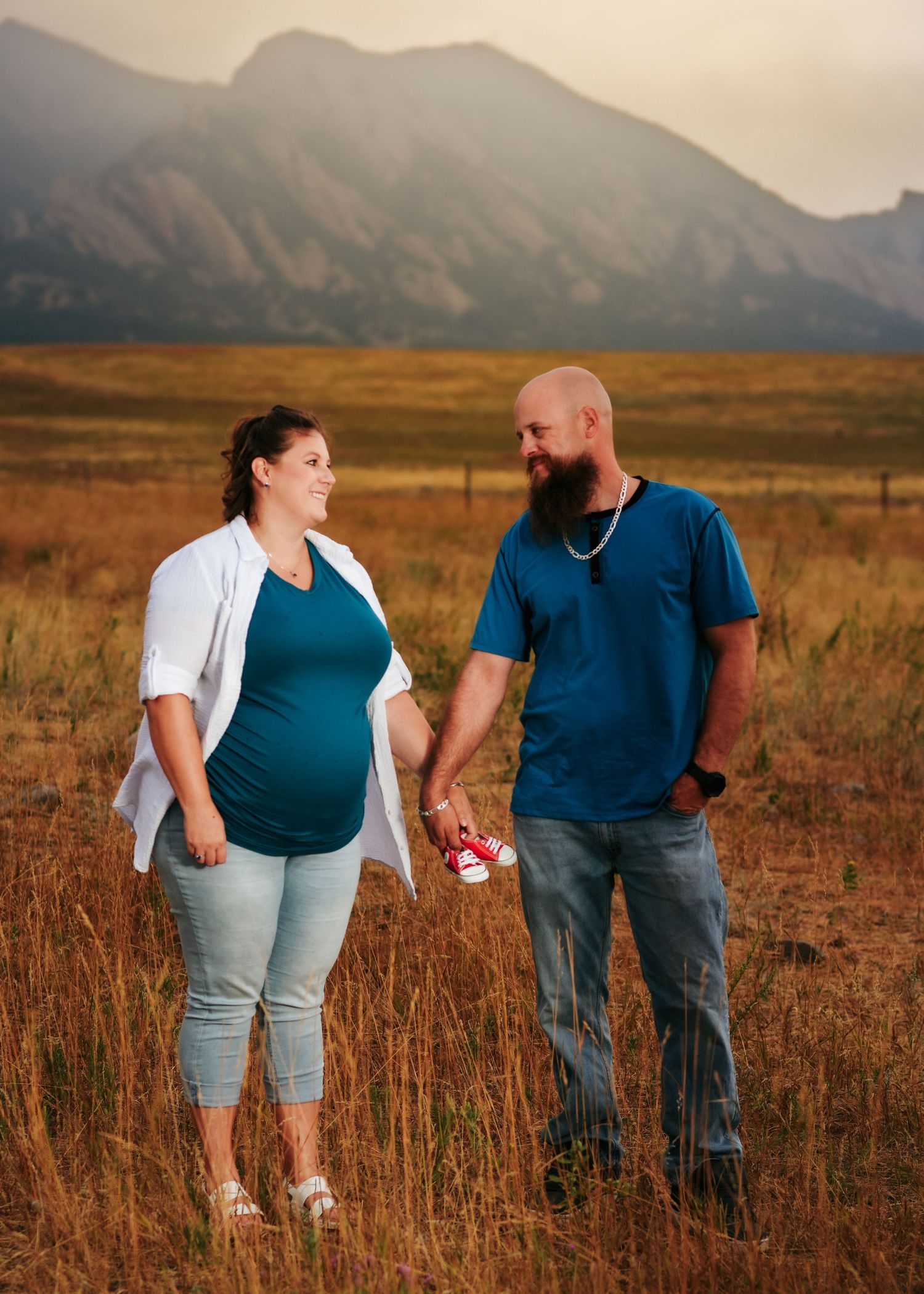 Pregnant couple holding tiny red shoes in a golden field with mountain backdrop during sunset maternity photo session.