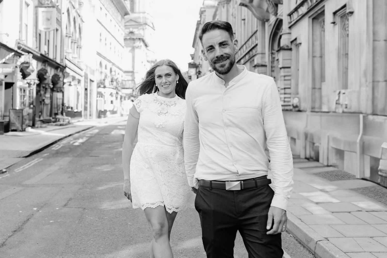 Black and white photo of a smiling couple walking together on a European city street, dressed in elegant casual attire.