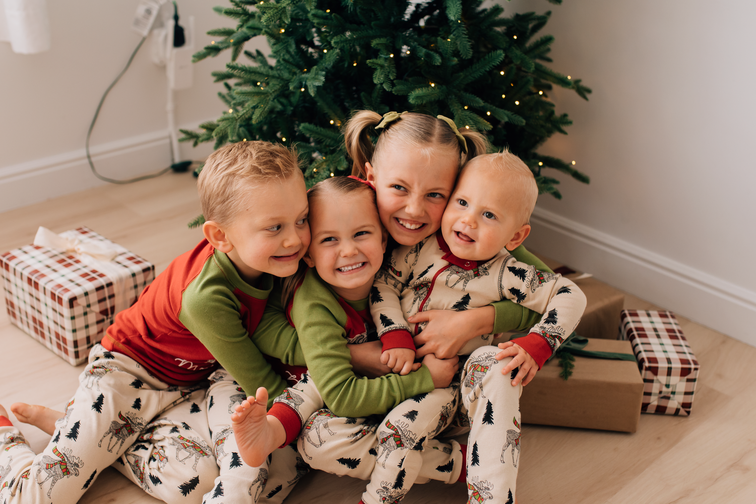 Four children in matching Christmas pajamas hugging near a decorated tree with wrapped gifts.